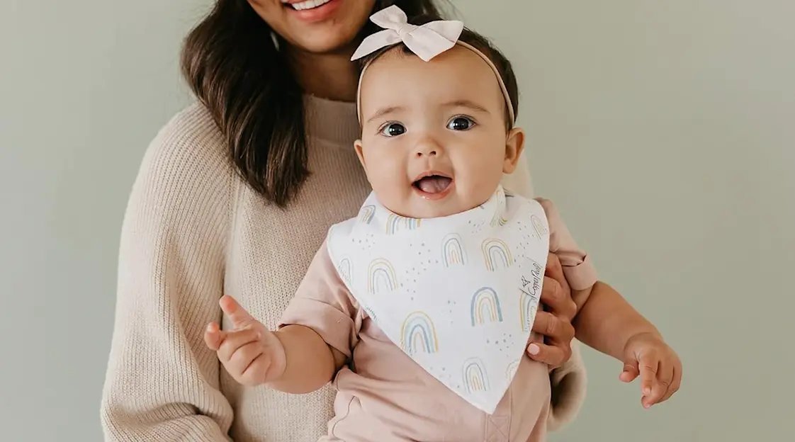 A cute baby wearing a soft absorbent bandana bib in a sunny kitchen.