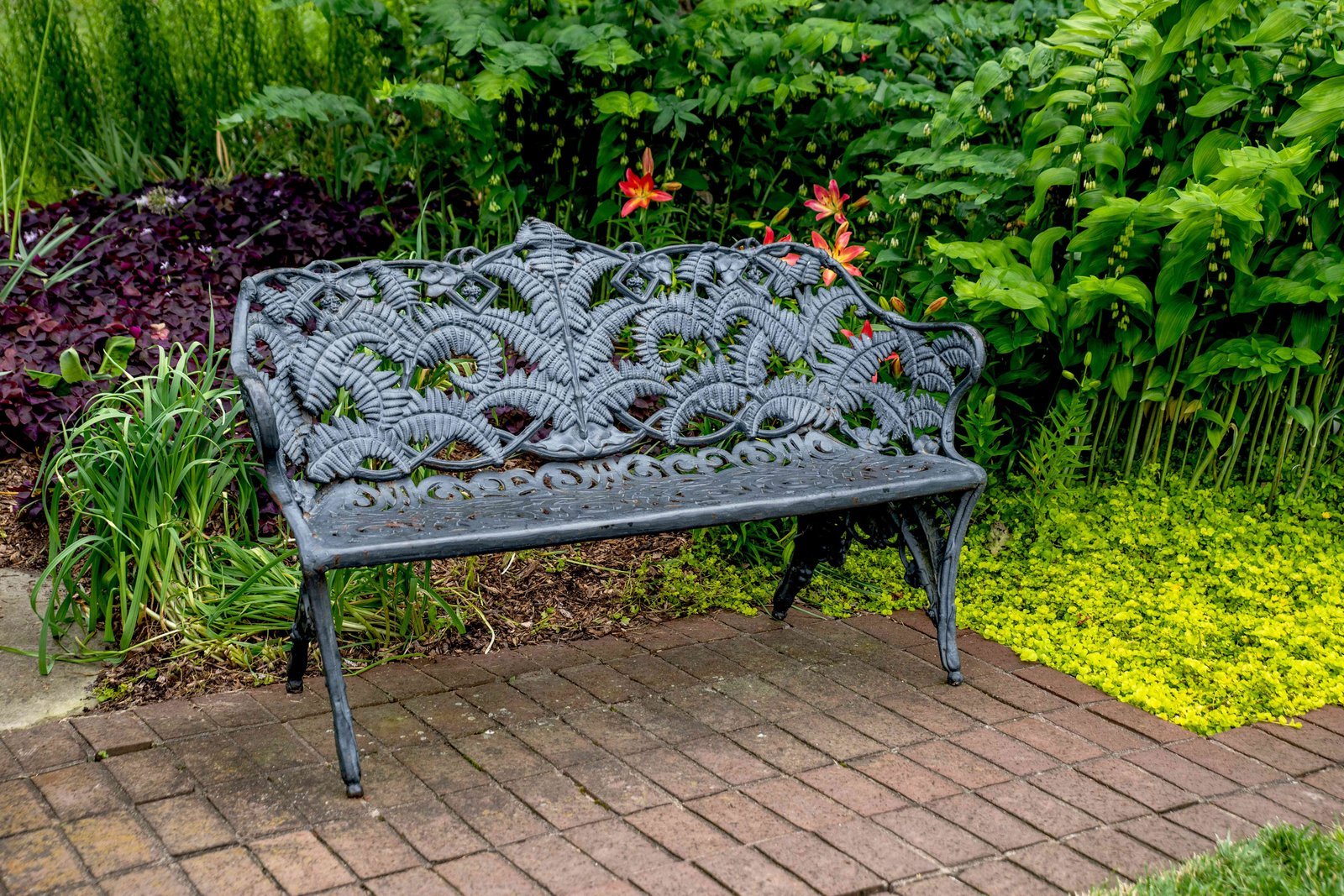 A modern teak garden bench sits on a lush lawn in a front yard.