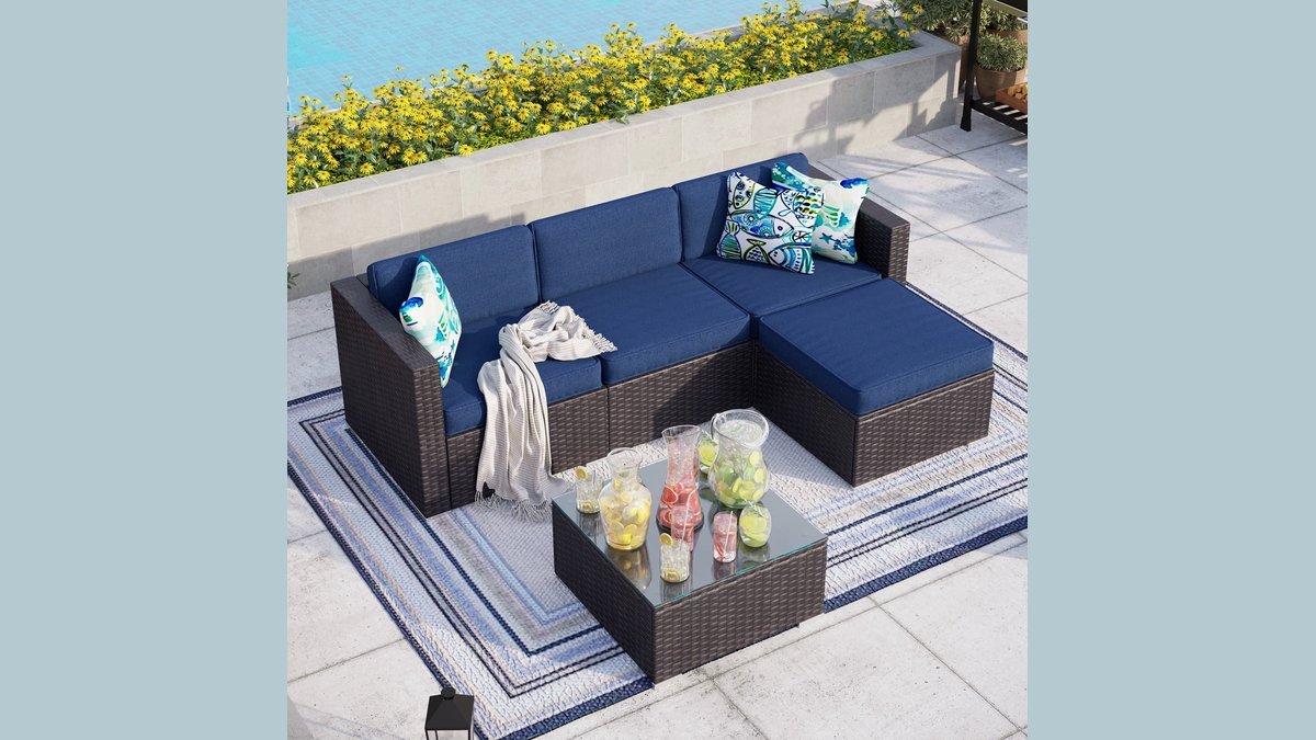A comfortable rattan outdoor sofa sits on a wooden patio surrounded by lush greenery.